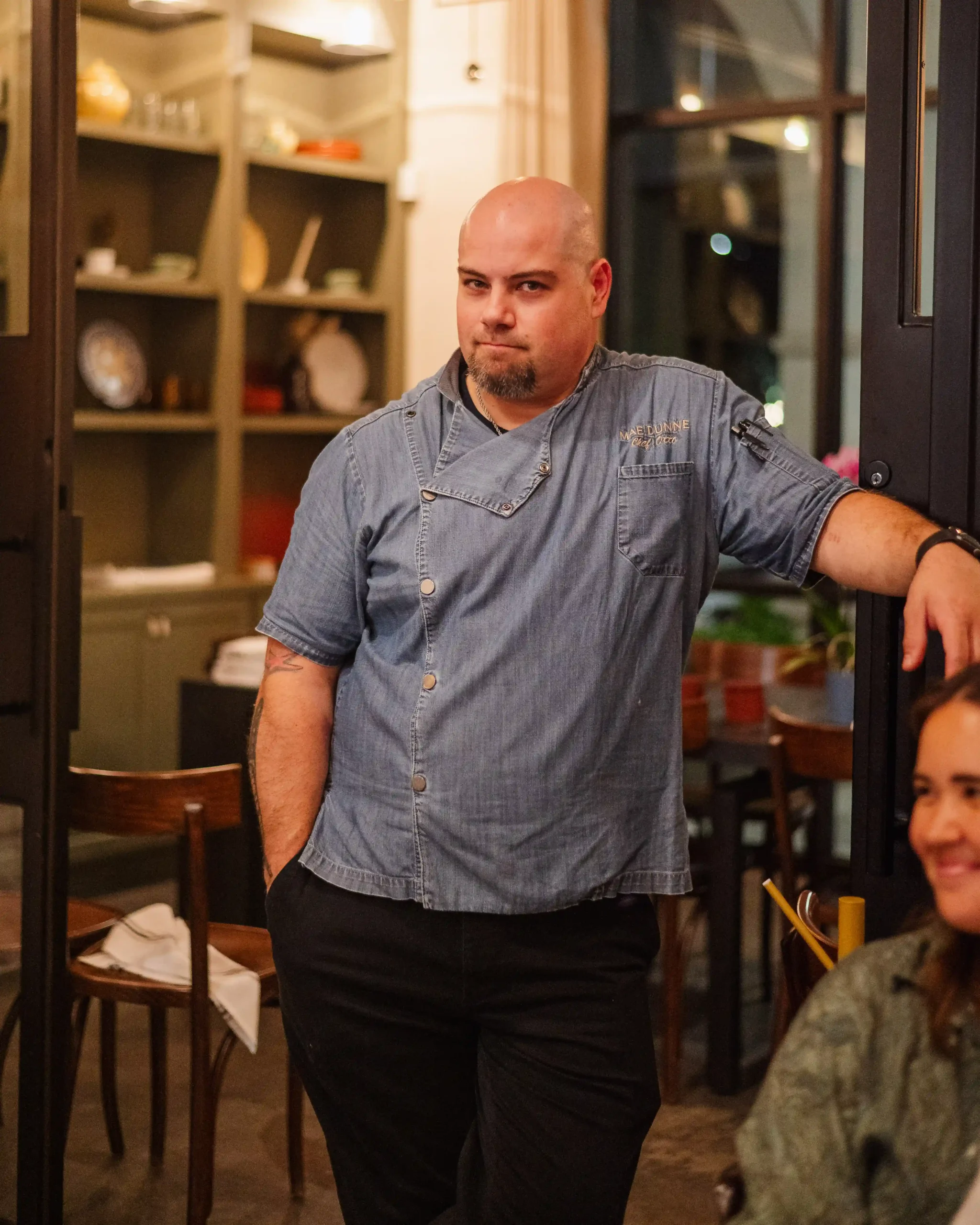 Chef in blue jacket standing inside restaurant with guests dining nearby.