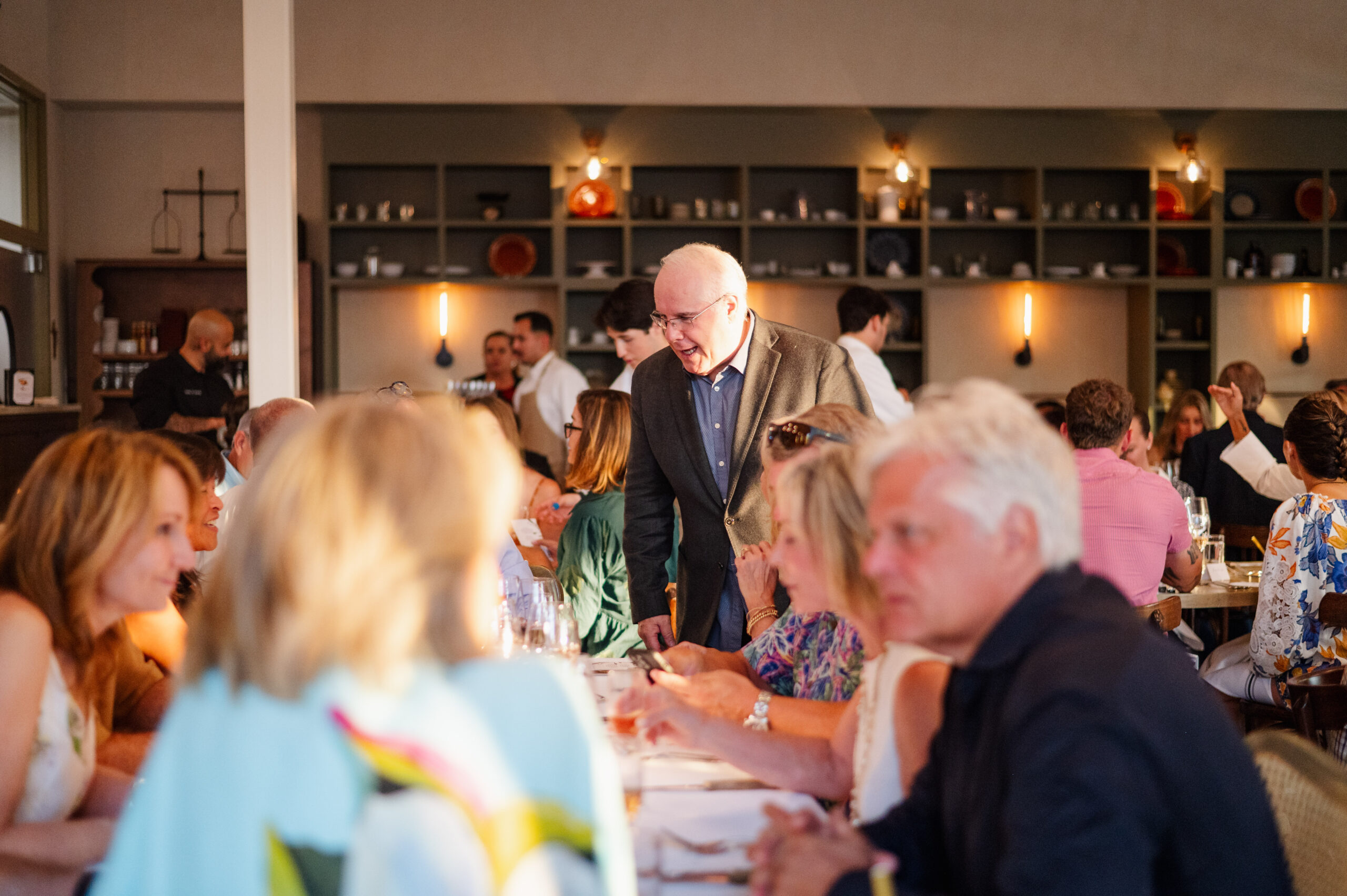 Elderly man engaging with diners during lively restaurant dinner event.